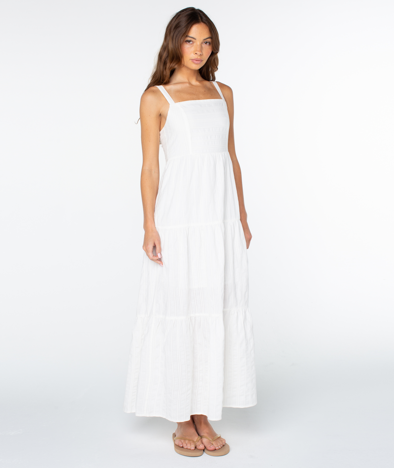 A woman with long brown hair wears the Roxy Clearwater Maxi Dress, featuring a sleeveless, white, square-neck design and beige sandals, standing against a plain white background.