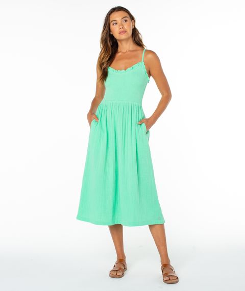 A woman stands with hands in pockets, wearing the Roxy Downriver Midi Dress—a sleeveless, coastal-style light green dress with thin straps and brown sandals—against a plain white background.