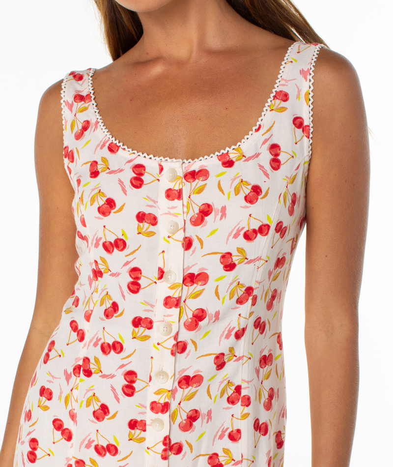A woman wears the Roxy Kayla Mini Dress, a sleeveless white summer dress with a cherry print, scoop neckline, lace trim, and front buttons. Only her upper half is visible.