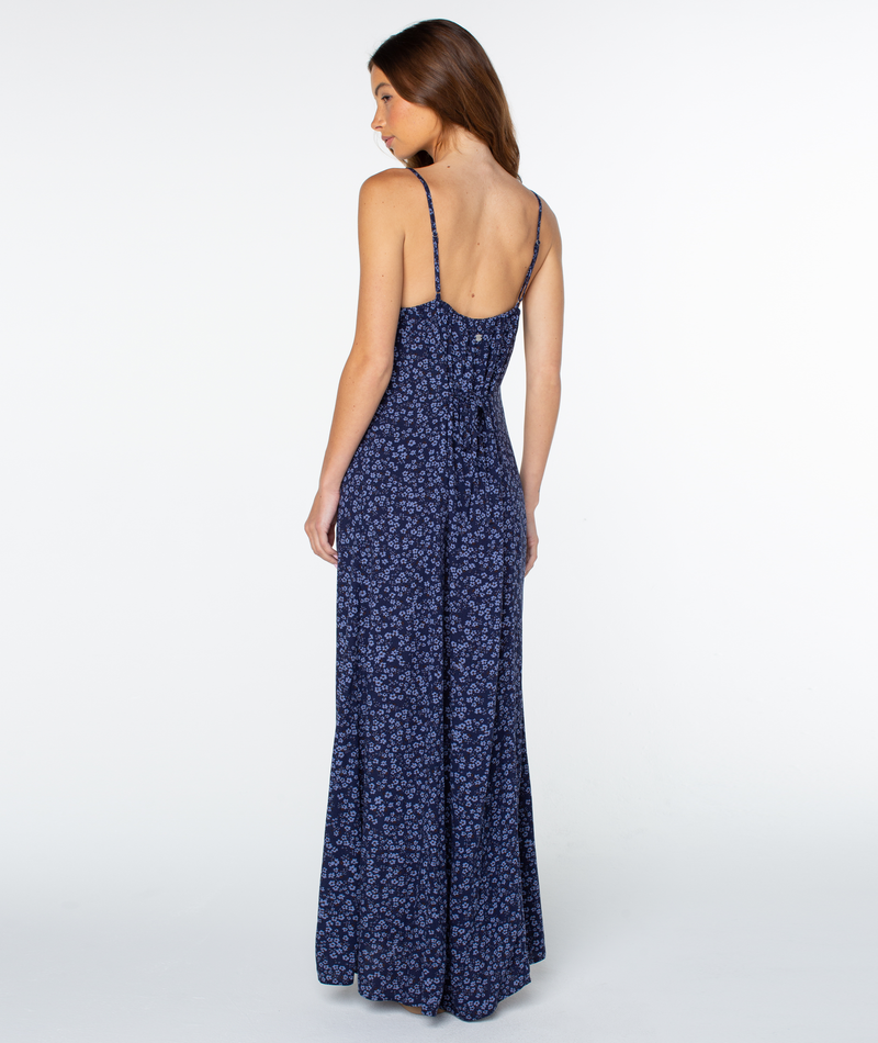 A woman with long brown hair wears the Roxy Skylar Maxi Dress in navy blue floral, featuring thin straps, an open back, and a square neckline, standing against a plain white background.