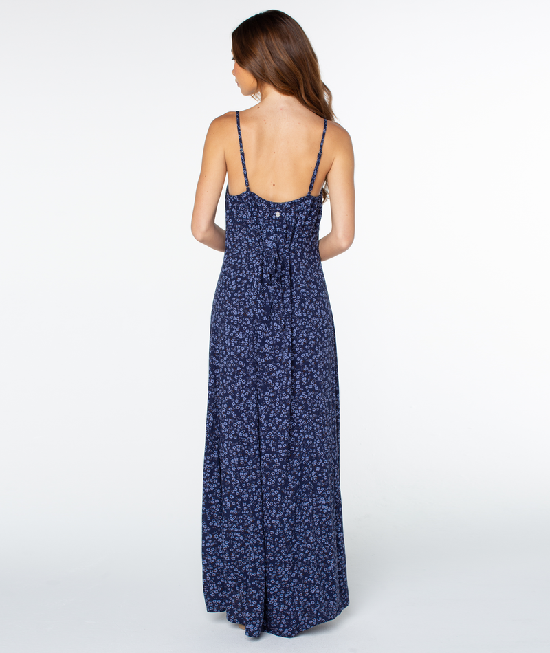 A woman with long brown hair, seen from behind, wears the Roxy Skylar Maxi Dress—a navy blue, sleeveless dress with a small floral print—standing on a plain white background.