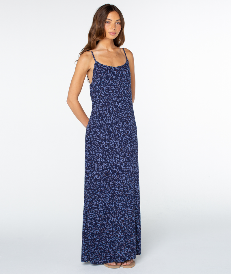 A woman wearing the Roxy Skylar Maxi Dress—a sleeveless, navy blue maxi with a small floral print and square neckline—stands against a white background, hands in pockets.