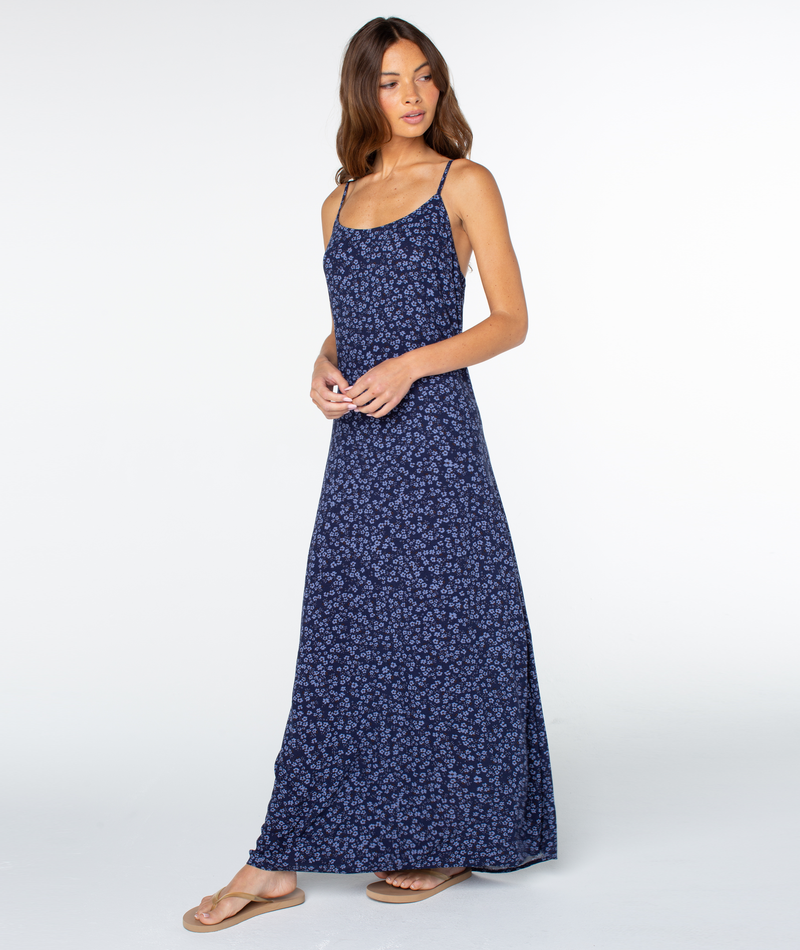 A woman in the Skylar Maxi Dress by Roxy—long, sleeveless, navy blue floral with a square neckline—stands in tan sandals against a white background, gazing to the side with a relaxed posture.