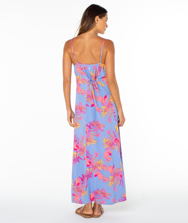 A woman faces away in the Roxy Skylar Maxi Dress—a long, light blue dress with vibrant florals and a back bow—ideal for tropical summer days. She pairs it with sandals, set against a plain white background.