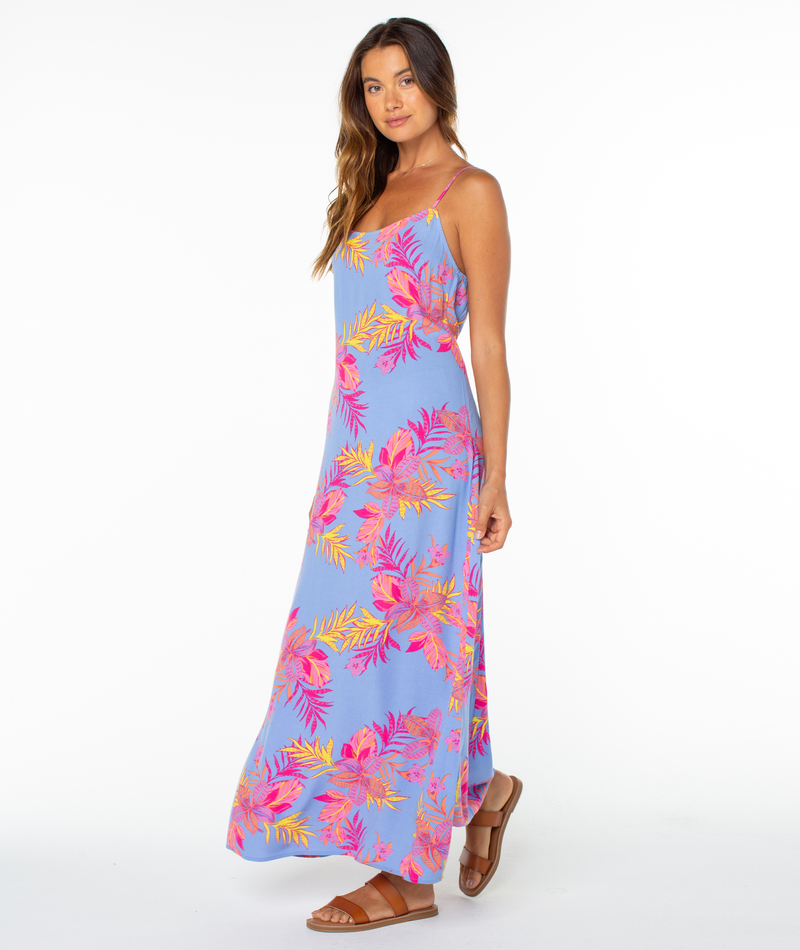 A woman in the Roxy Skylar Maxi Dress—a long, sleeveless blue dress with bright florals and tropical leaf patterns—pairs brown sandals and loose hair, posing against a white background for a breezy summer look.