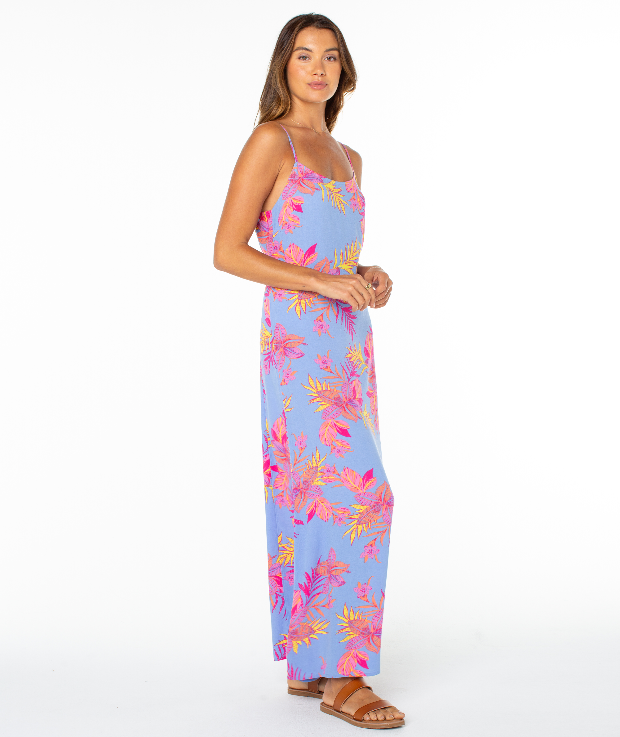 A woman poses sideways in a sleeveless, light blue Roxy Skylar Maxi Dress with bright florals, paired with brown sandals against a plain white background.