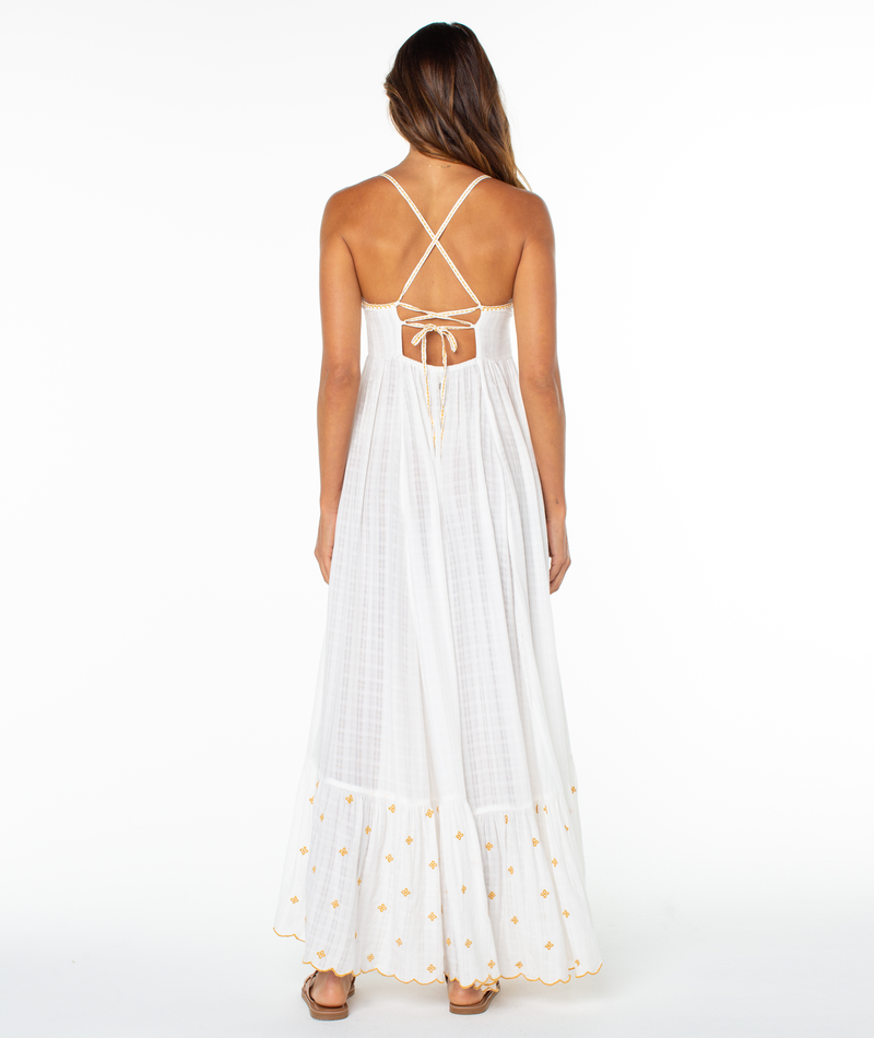 A woman with long, wavy hair stands barefoot on a white background, wearing the Roxy Sunburst Maxi Dress—a white summer dress featuring a crisscross tie-back and golden embroidery near the hem.