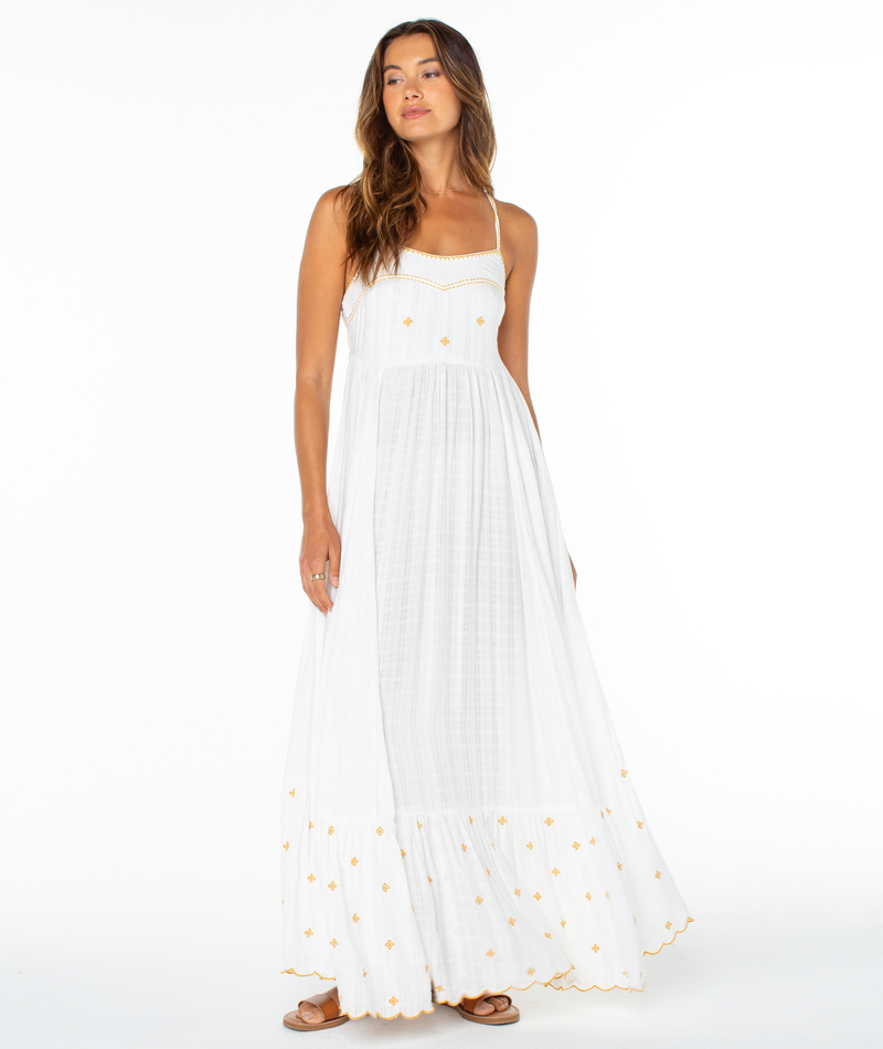 A woman with long, wavy brown hair and a relaxed expression stands barefoot against a white background wearing the Roxy Sunburst Maxi Dress, a long, sleeveless white summer dress with gold embroidery along the top and hem.