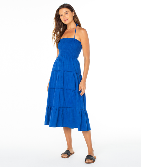 A woman with long brown hair stands against a white background wearing the Roxy Ellie Dress, a sleeveless, halter-neck style in tiered royal blue, paired with black slide sandals. She appears relaxed.