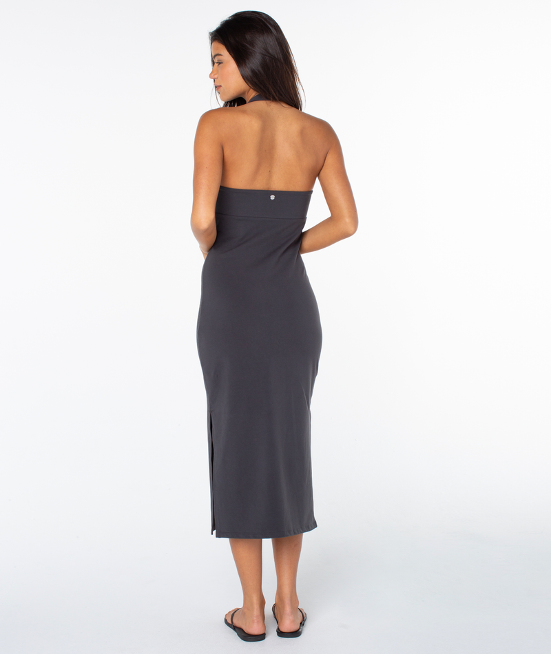 A woman faces away from the camera in the Roxy South Shore Midi Dress—a strapless, charcoal gray, ankle-length dress with a side slit—styled with black flip-flops against a plain white background.