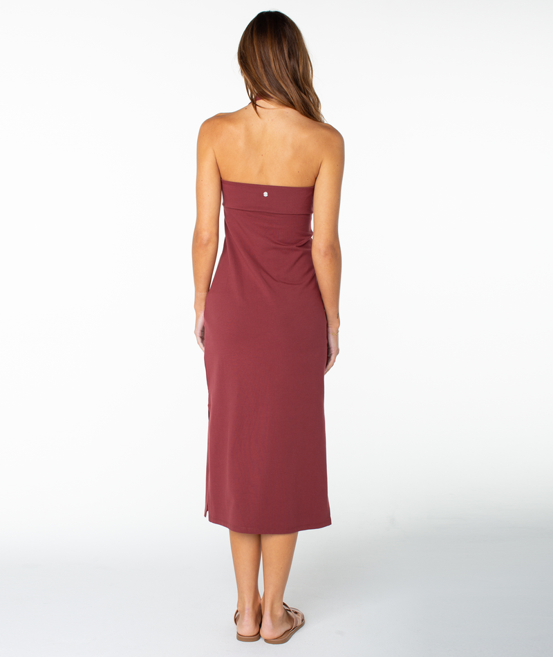 A woman stands barefoot, facing away, wearing the South Shore Midi Dress by Roxy—a strapless, ankle-length maroon piece. Her brown hair is loose against a plain white background for an effortless off-duty look.
