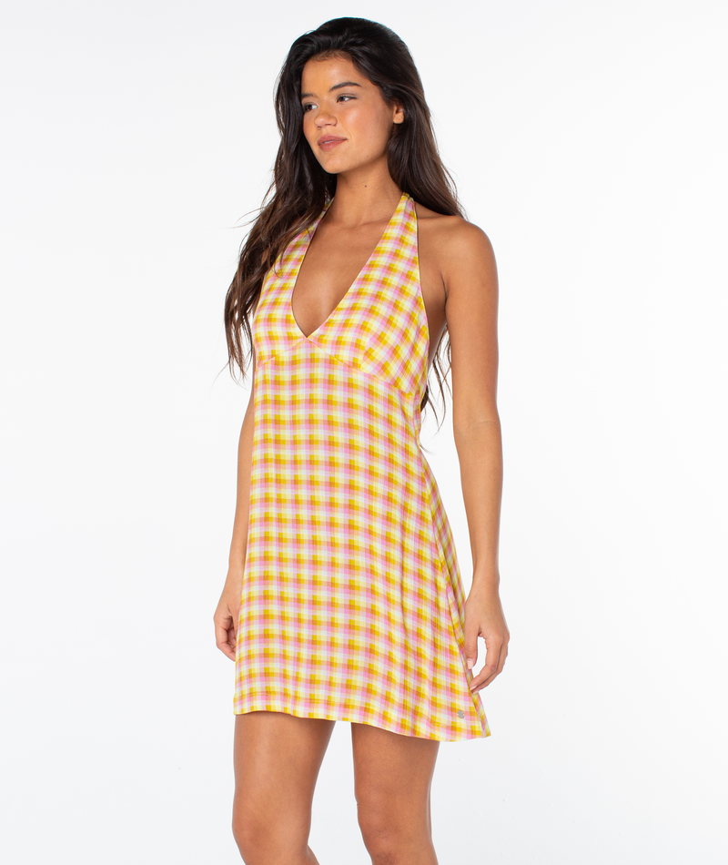 A woman with long dark hair models the Roxy Beach Day Mini Dress, a sleeveless yellow and pink plaid halter style with a deep V-neck, standing against a plain white background.