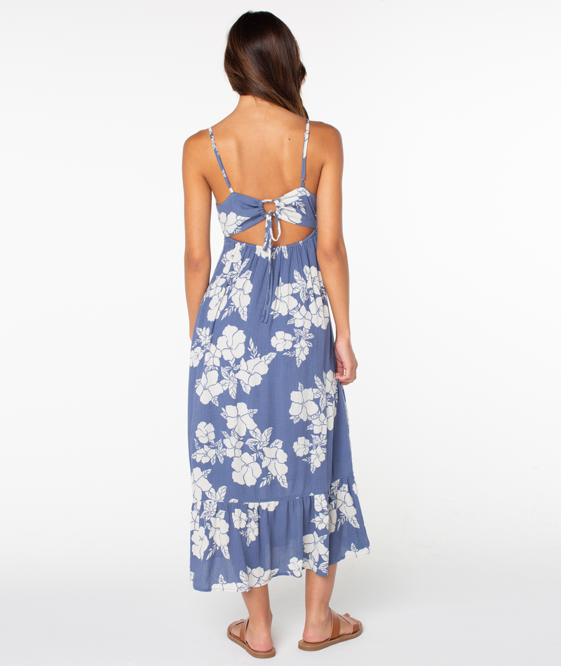 A woman with long brown hair wears the Roxy Warm Rays Midi Dress, featuring a blue and white hibiscus print, spaghetti straps, open back, and bow tie. She stands barefoot facing away against a white background for tropical vibes.