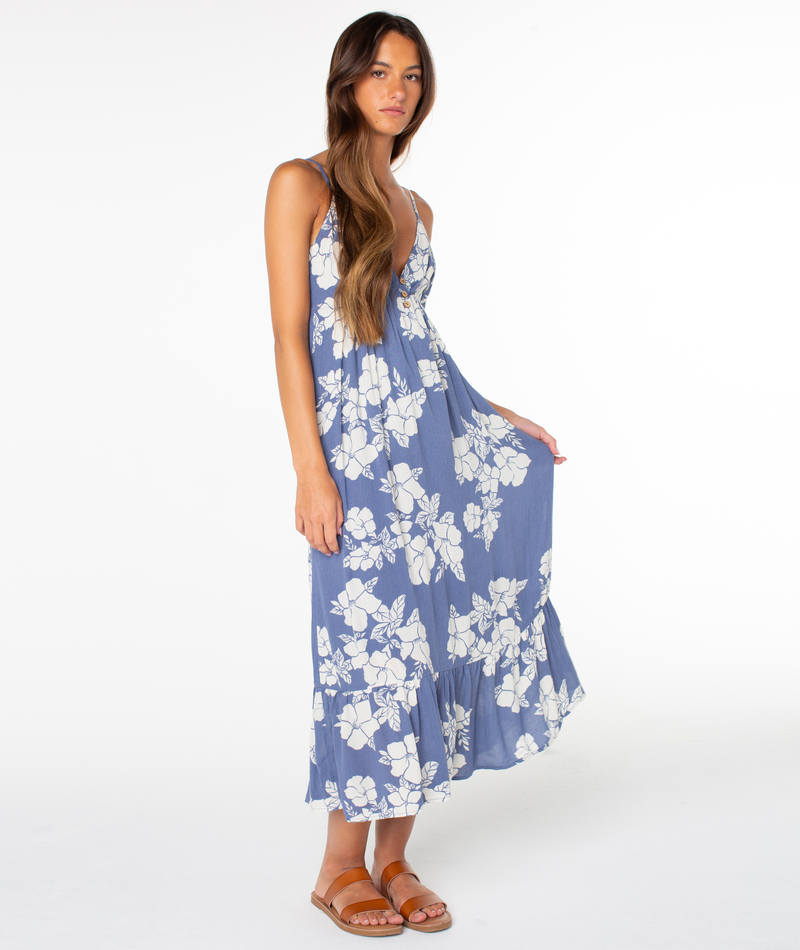 A woman with long brown hair models the Roxy Warm Rays Midi Dress, a blue sleeveless hibiscus print style, paired with brown sandals. She stands against a white background, holding her dress and exuding tropical vibes.