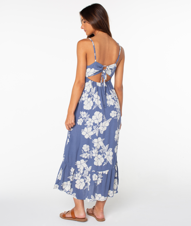 A woman stands turned away in the Roxy Warm Rays Midi Dress—a blue, white hibiscus-print dress with an open back and tie detail. She has long brown hair, wears brown sandals, and radiates tropical vibes against a plain white background.