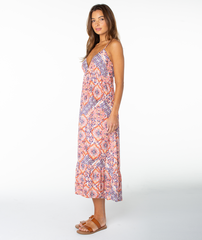 A woman stands sideways in the Roxy Warm Rays Midi Dress, a sleeveless, long hibiscus print sundress in peach, blue, and white, paired with brown flat sandals against a white background for fresh tropical vibes.