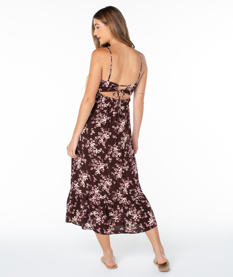A woman with long hair wears the Roxy Warm Rays Midi Dress—sleeveless, backless, with a pink floral maroon print—and sandals, standing and glancing over her shoulder against a white background for tropical vibes.