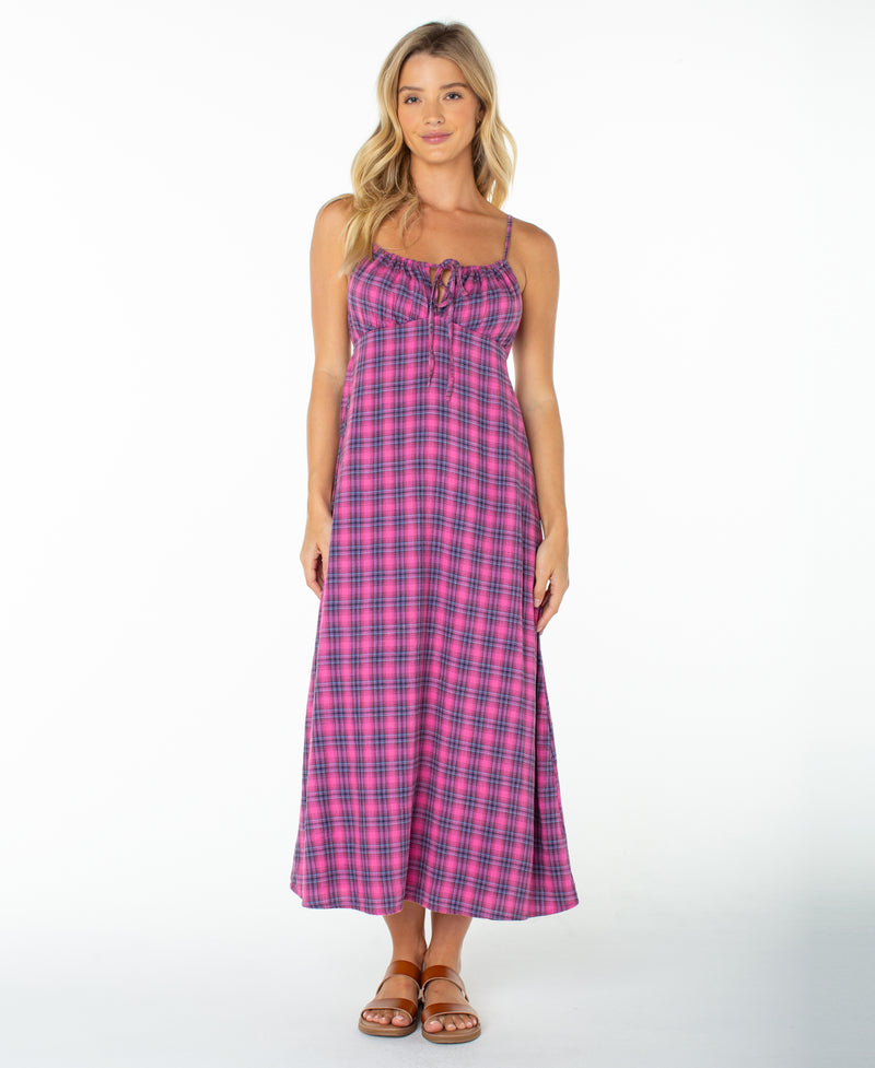 A woman wearing the Roxy Late Night Midi Dress in Naval Academy Sunburst Plaid—a long, sleeveless midi dress with spaghetti straps and a ruched drawstring neckline—paired with brown sandals, stands against a plain white background.
