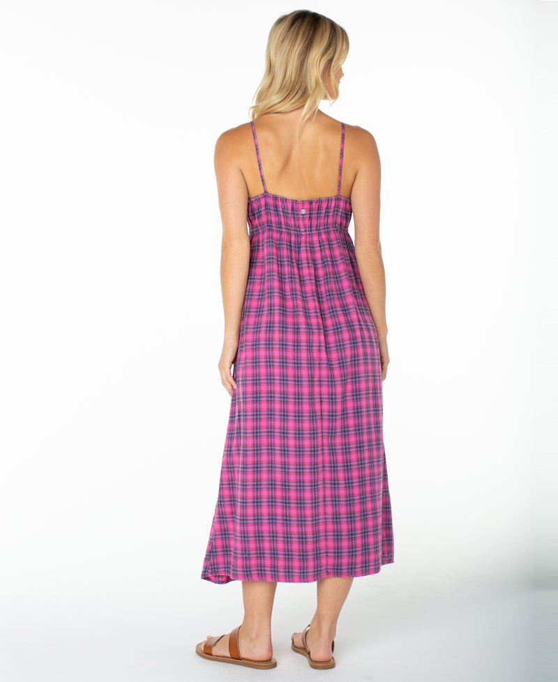 A woman with blonde hair, wearing the Roxy Late Night Midi Dress in a pink and blue plaid with a ruched drawstring neckline and brown sandals, stands facing away against a plain white background. This dress is perfect for any beachside occasion.