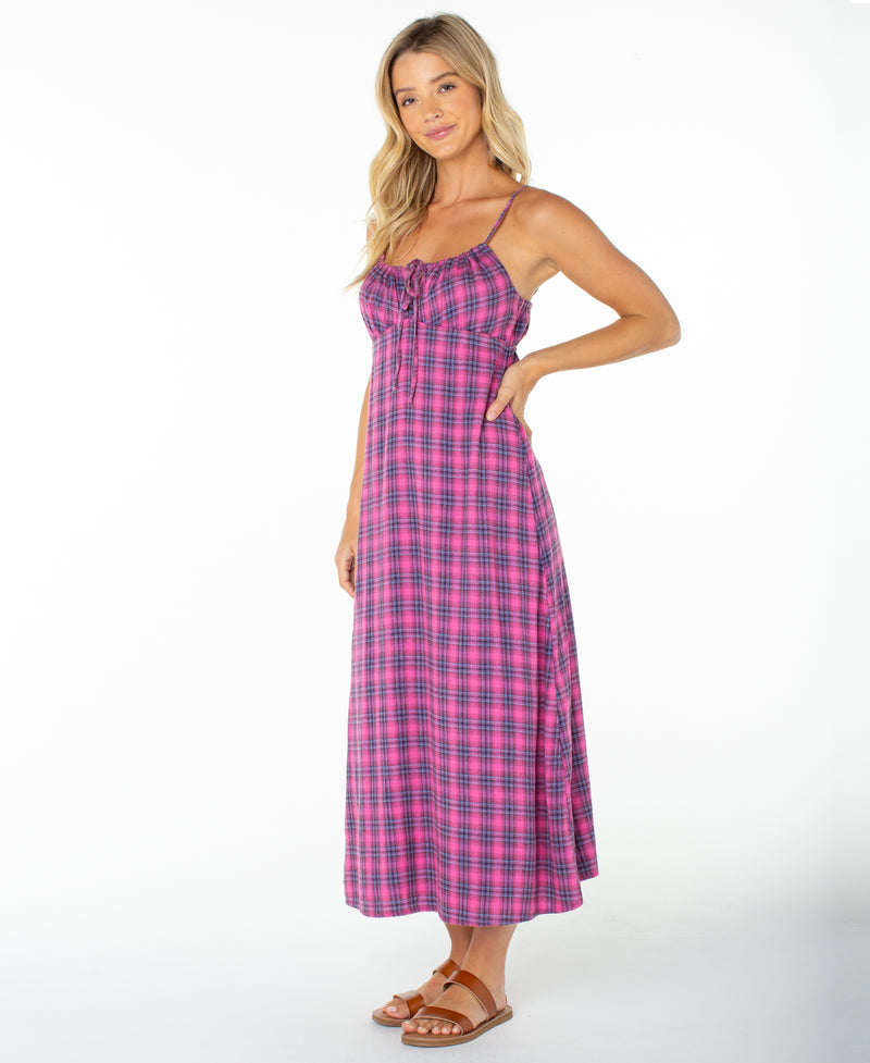 A woman stands in the Roxy Late Night Midi Dress, featuring a pink and blue plaid pattern, spaghetti straps, and a ruched drawstring neckline. Paired with brown sandals against a white background, this sleeveless dress is perfect for any beachside occasion.