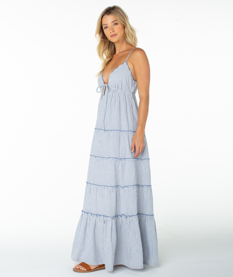 A woman models the Roxy Starlight Maxi Dress—a light blue and white striped summer dress with thin straps and a tiered skirt—paired with brown sandals, posing against a plain white background.