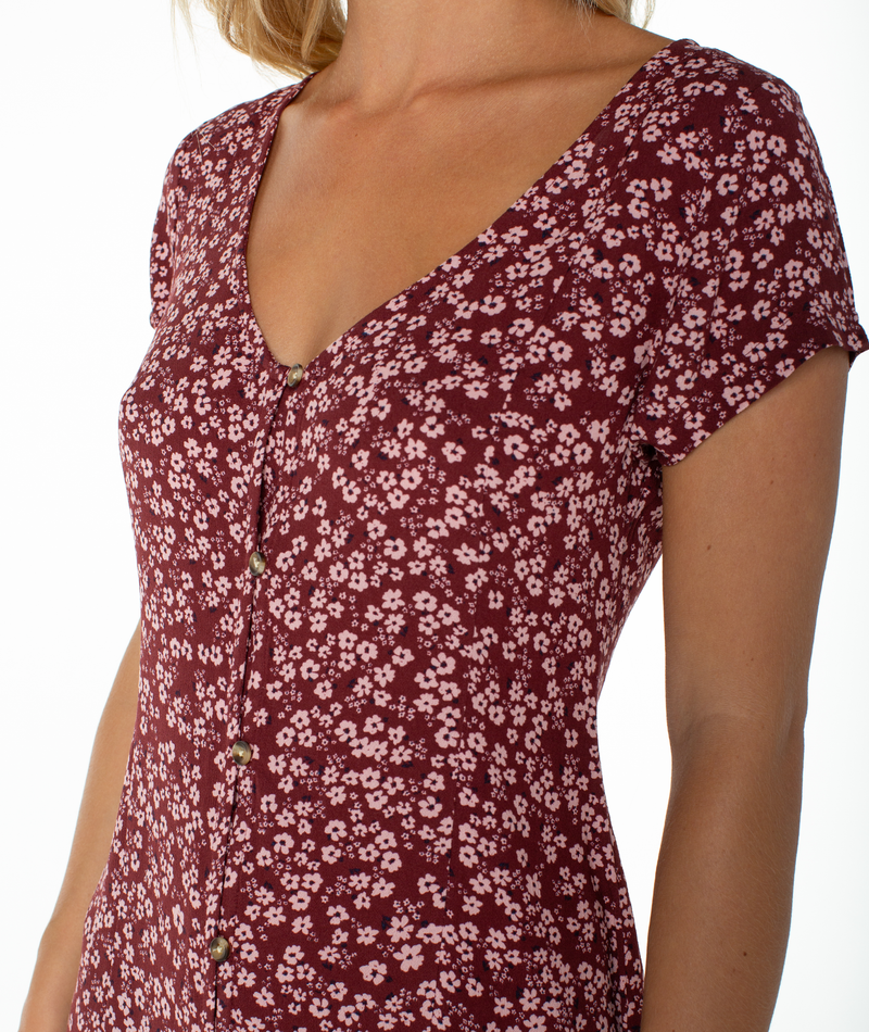 A person wearing the Roxy Cool Breeze Mini Dress, a short-sleeved burgundy dress with a pink floral pattern and front buttons. Featuring a subtle scoop neckline, the image is cropped at the shoulders and midsection.