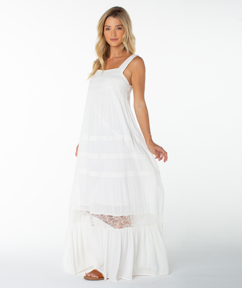 A woman in the Roxy Summer Wind Maxi Dress—a long, sleeveless white dress with lace detail and a square neckline—stands against a plain white background, looking at the camera with a slight smile while gently holding her dress.