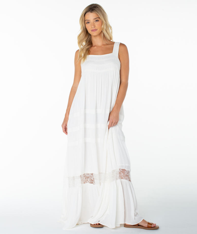 A woman with long blonde hair models the Roxy Summer Wind Maxi Dress, a sleeveless, flowing maxi with lace detail and a square neckline, standing against a white background and gazing at the camera with a relaxed look.
