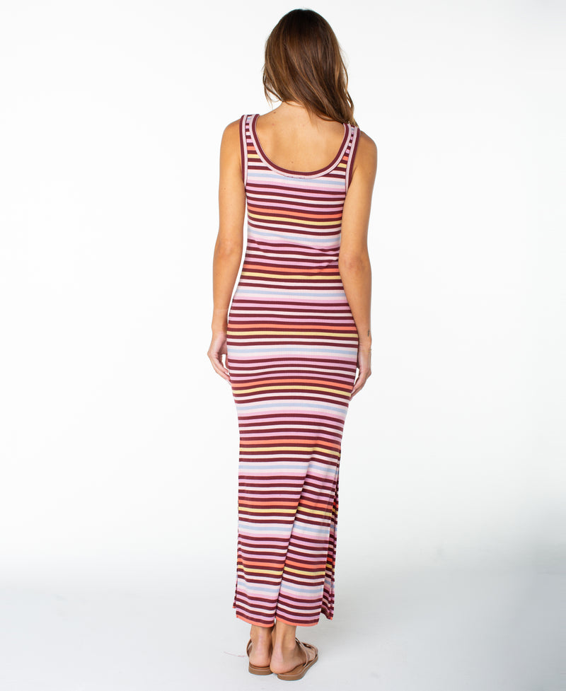 A woman stands facing away, wearing the Roxy Balboa Maxi Dress—a sleeveless style featuring horizontal pink, red, and white stripes. Crafted from a soft cotton blend, the stripe maxi dress falls to her ankles. She wears sandals, and the scene is set against a plain white background.