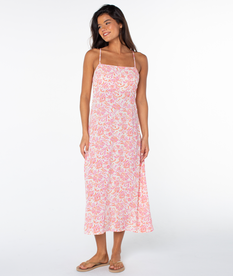 A woman with long dark hair smiles, looking to the side, wearing a sleeveless pink and white Roxy Balmy Sunset Dress and flip-flops, posing against a plain white background.