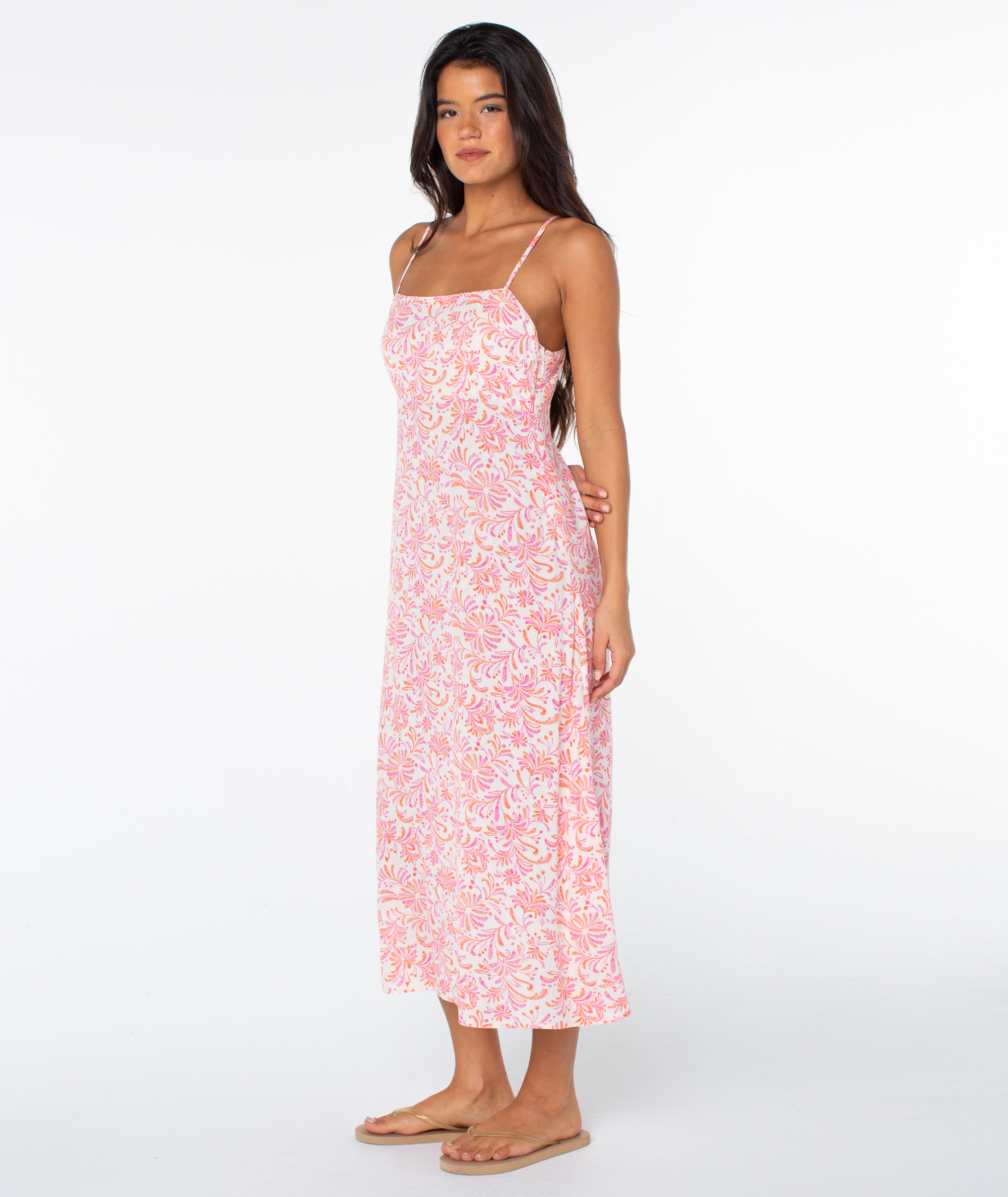 A woman stands barefoot on a white background wearing the Roxy Balmy Sunset Dress, a sleeveless, ankle-length tropical floral maxi dress with thin straps, looking slightly to the side.