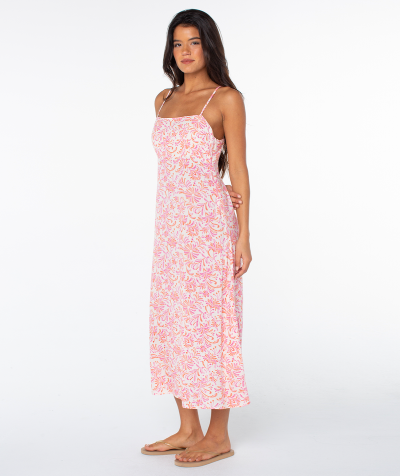 A woman stands barefoot on a white background wearing the Roxy Balmy Sunset Dress, a sleeveless, ankle-length tropical floral maxi dress with thin straps, looking slightly to the side.