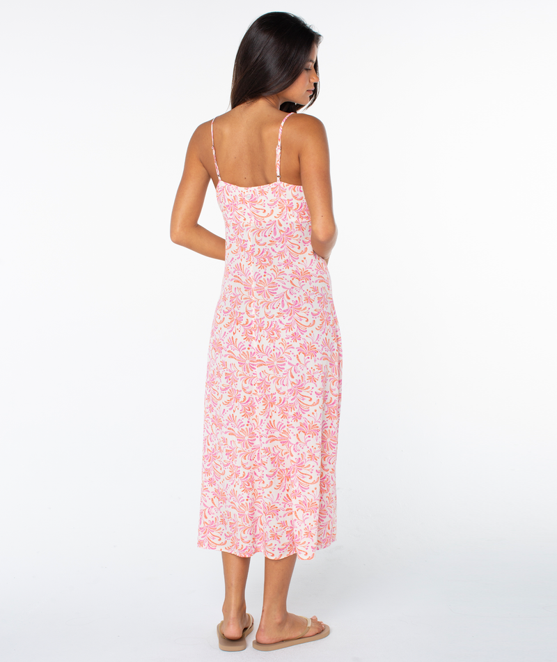 A woman with long brown hair, wearing the Roxy Balmy Sunset Dress—sleeveless with pink and white tropical florals—and beige sandals, stands facing away against a plain white background.