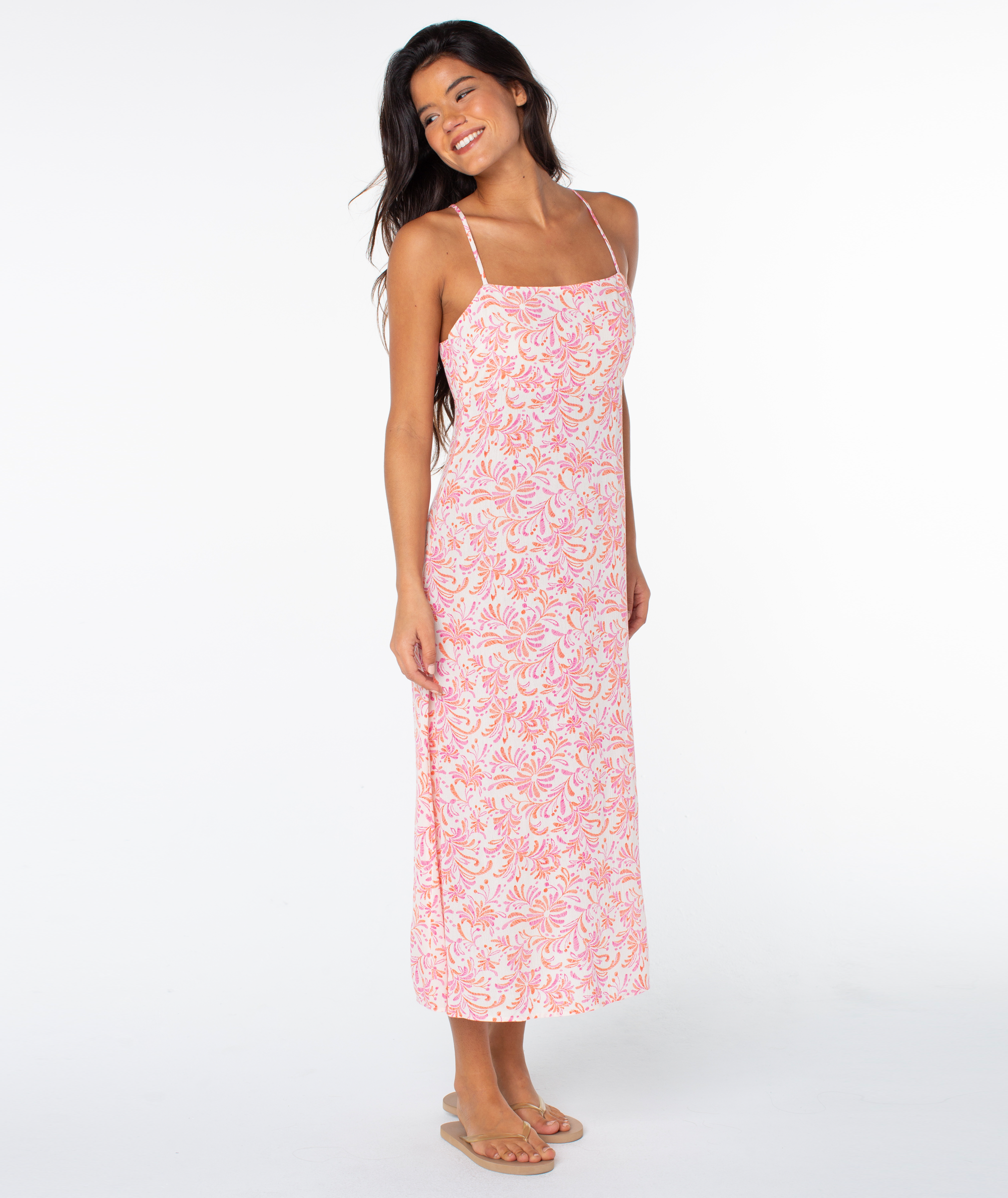 A woman in a sleeveless, ankle-length Roxy Balmy Sunset Dress with thin straps and flip-flops stands smiling against a plain white background.