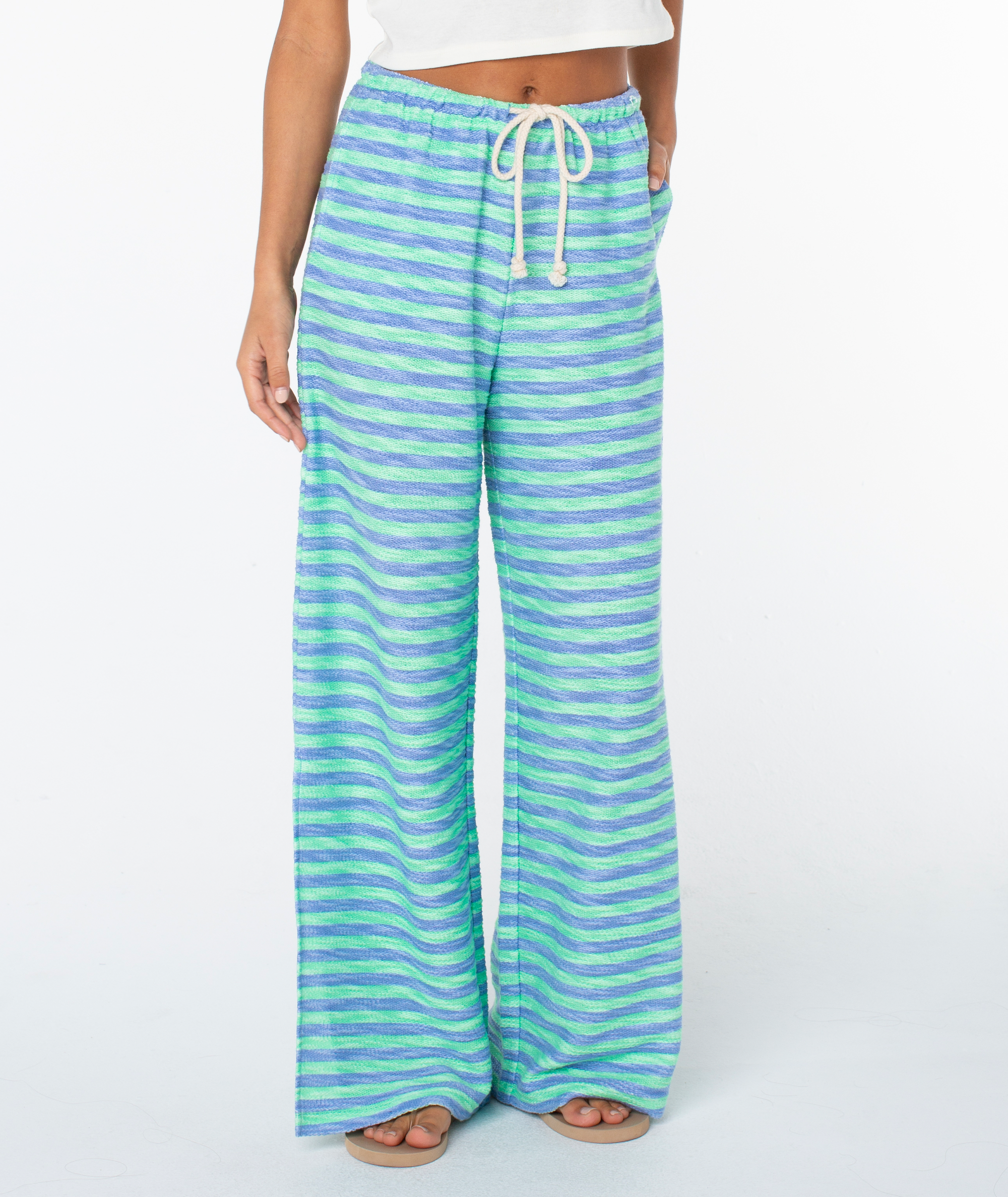 A person wears the Roxy Beach Breeze Pant, featuring wide legs, a high waist, blue-green-purple horizontal stripes, an adjustable drawstring waist, and pockets, paired with a white cropped top.