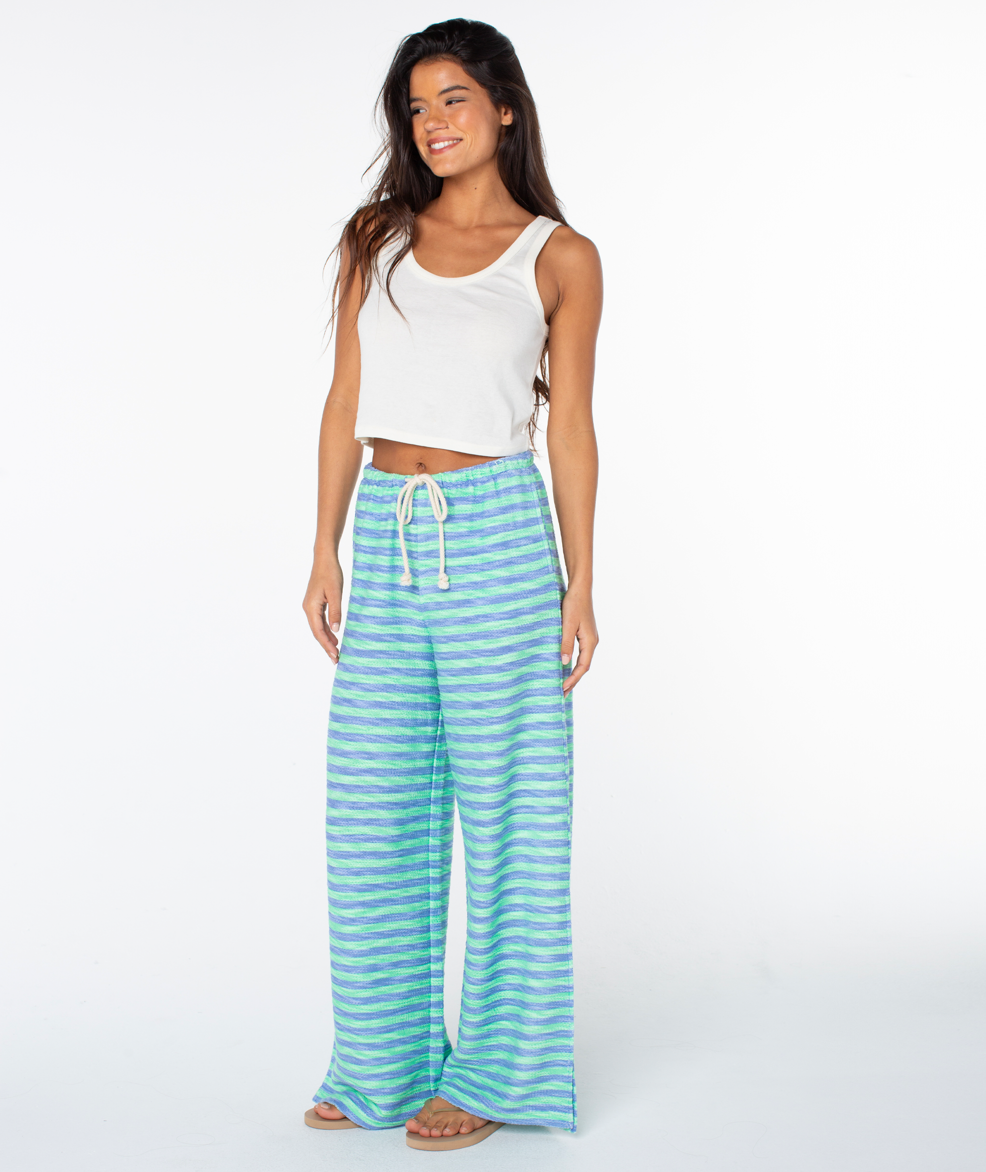 A woman smiles in a white sleeveless crop top and Roxy Beach Breeze Pant—blue and green striped, loose, wide-legged cotton blend pants with a drawstring waist—paired with sandals against a plain white background.