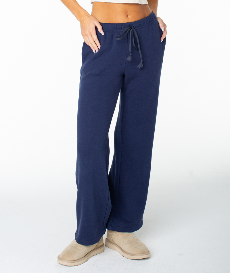 Person wearing navy blue Roxy Good Times Pant sweats and tan slippers, with hand resting on hip, shown from torso down against a plain white background—pairs perfectly with the matching Dream Thing top.