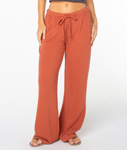 A person is shown from the waist down wearing Roxy’s Good Times Pant in Chutney Solid—a loose, rust-colored style with a relaxed fit, sweats-inspired comfort, convenient pockets, and a drawstring waist. They’ve paired the pants with sandals and are standing against a white background.