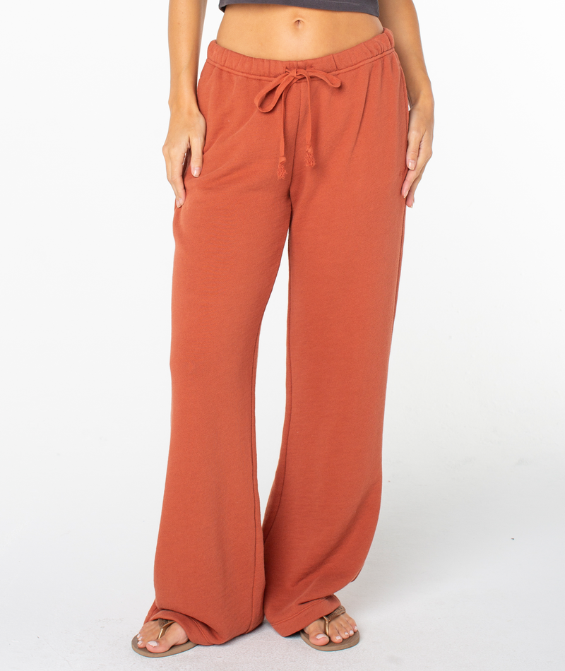 A person is shown from the waist down wearing Roxy’s Good Times Pant in Chutney Solid—a loose, rust-colored style with a relaxed fit, sweats-inspired comfort, convenient pockets, and a drawstring waist. They’ve paired the pants with sandals and are standing against a white background.