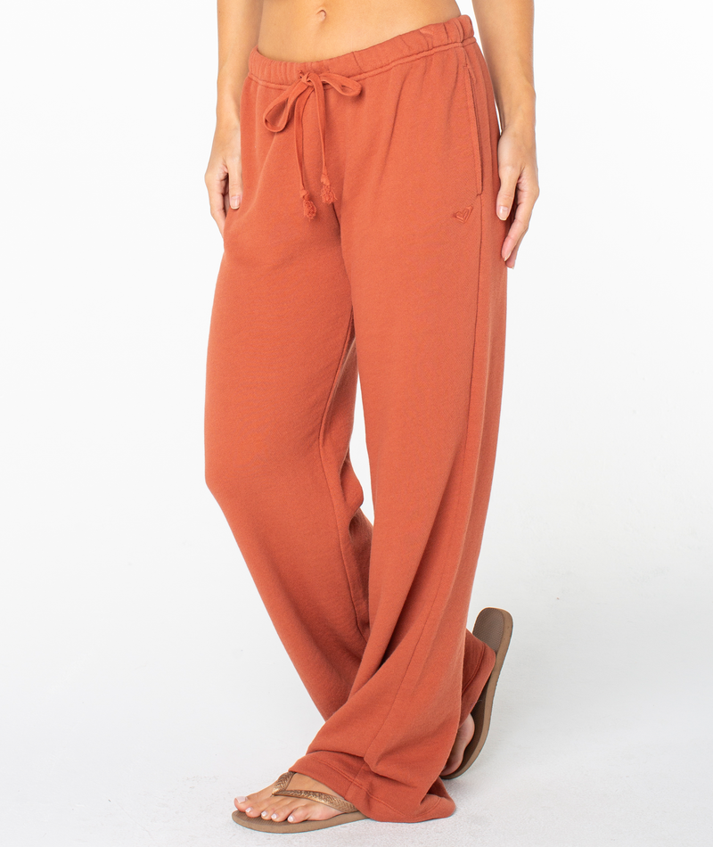 A person models the Roxy Good Times Pant in a relaxed, rust color with a flattering silhouette and sandals, standing against a plain white background. Only the lower half of their body is visible.