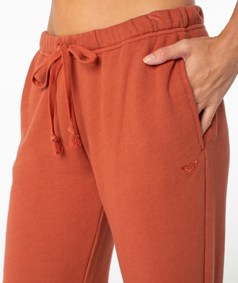 A close-up of a person wearing the Roxy Good Times Pant in burnt orange, featuring a drawstring waist, one hand casually placed in the pocket, and a small embroidered heart logo on the hip. Crafted for a flattering fit and all-day comfort.