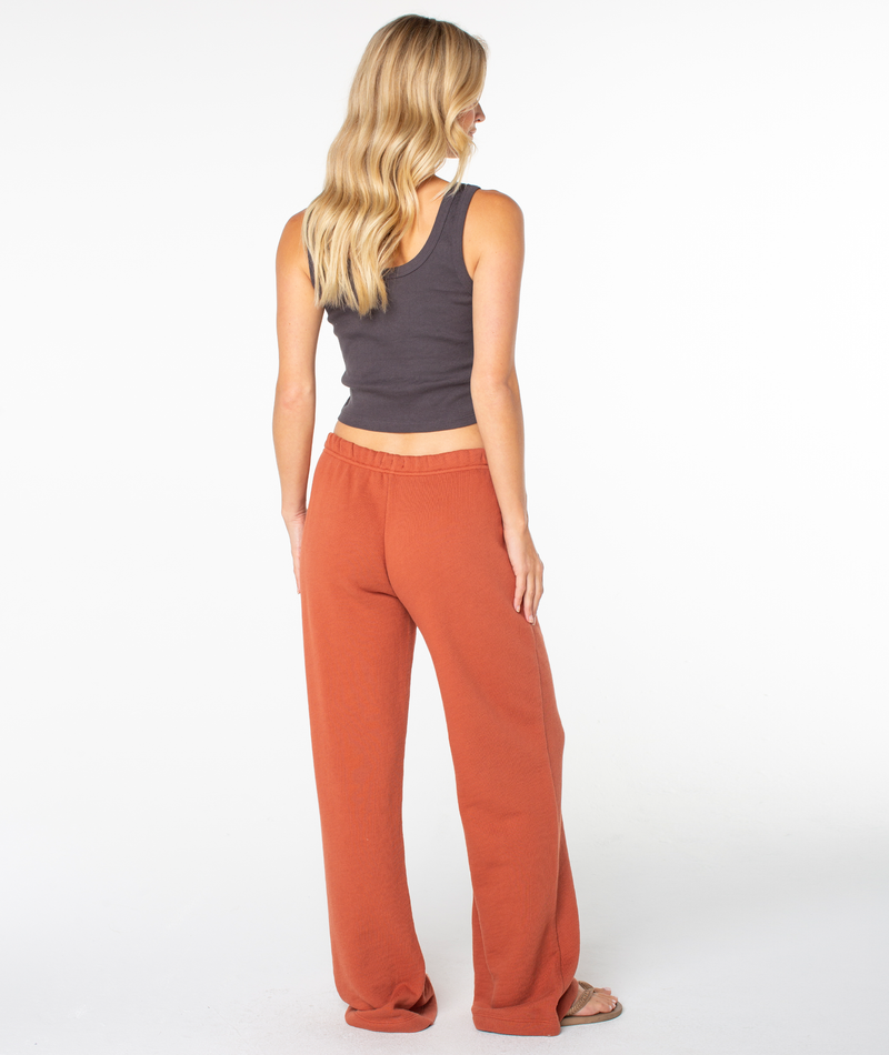 A woman with long blonde hair, wearing a dark gray tank top and loose orange Roxy Good Times Pant sweats that create a flattering shape, stands barefoot facing away in a plain studio setting.