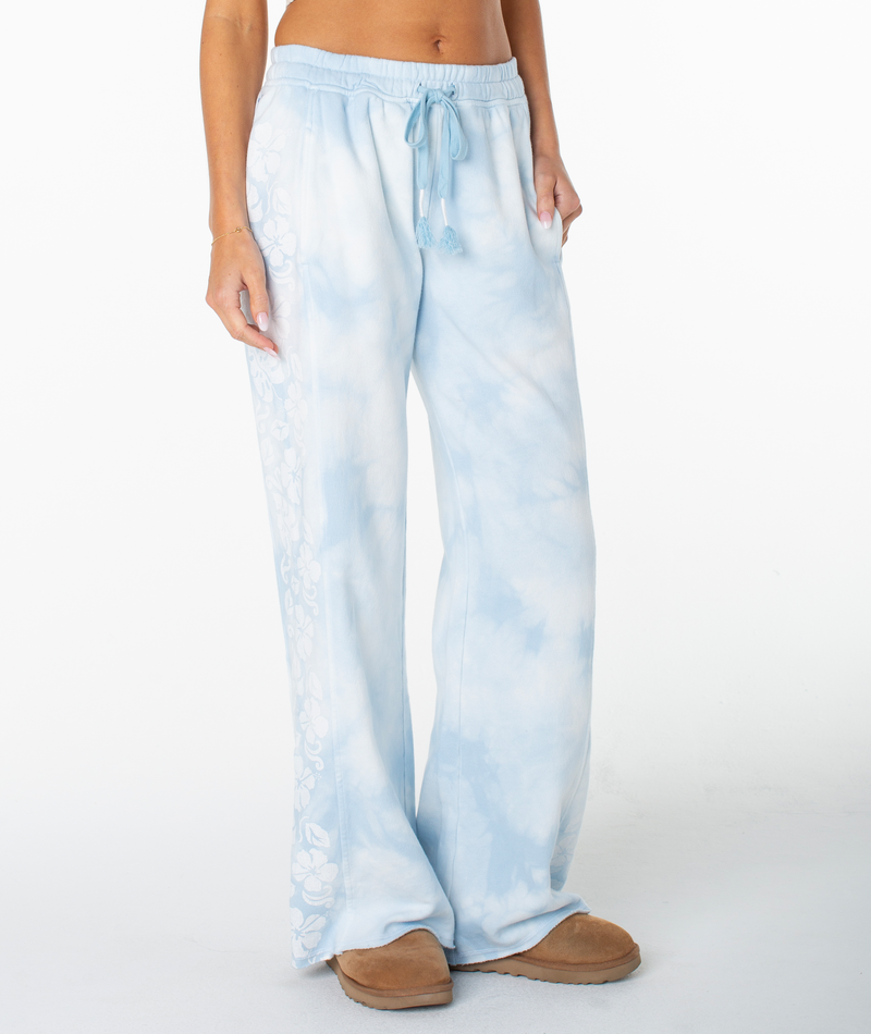 A person is lounging in Roxy’s Fringing Reefs Pant in Cerulean Tie Dye, featuring a drawstring waist and standing with one hand in a pocket against a white background, paired with brown slippers.