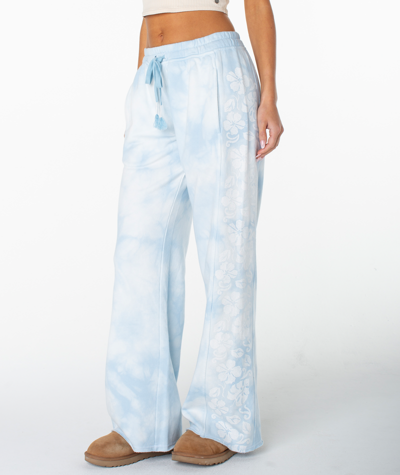 A woman wearing Roxy’s Fringing Reefs Pant—light blue tie-dye wide-leg pants featuring a white floral print along the sides—paired with tan slip-on shoes and a cropped white top, stands against a white background for an effortlessly lounging look.