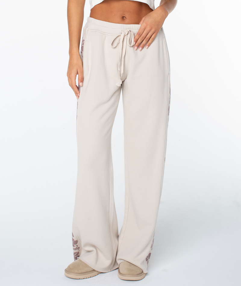 A person wears the Fringing Reefs Pant by Roxy—beige lounging pants with leaf patterns on the sides—paired with matching beige slippers, midriff and hands visible, set against a plain white background.