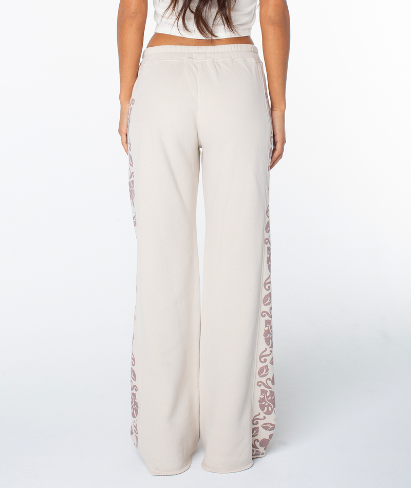 A woman faces away, wearing Roxy’s Fringing Reefs Pant—high-waisted, beige lounging pants with floral side seams—paired with a cropped white top.