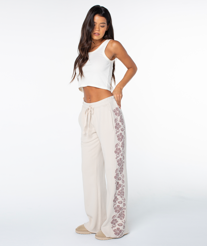 Woman wears Roxy Fringing Reefs Pant in Pumice Stone Fringing Reefs with a white tank top, standing against a plain white background, looking down with one hand on her hip.