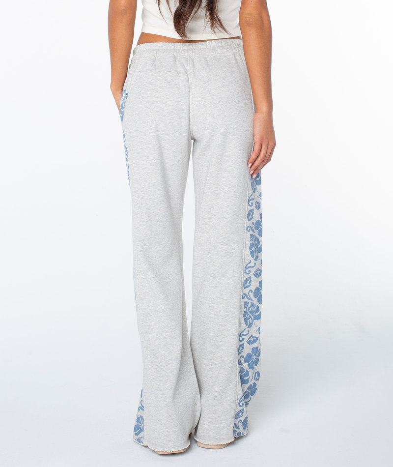 A woman stands with her back to the camera in the Roxy Fringing Reefs Pant, featuring light gray fabric and blue floral side panels, paired with a white top against a plain white background.