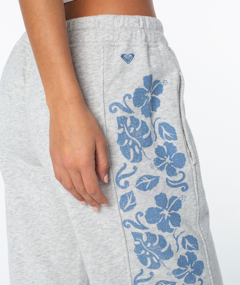 A person models the Roxy Fringing Reefs Pant—light gray with blue floral patterns and a small heart logo—showing their hand, hip, and upper leg against a plain white background.