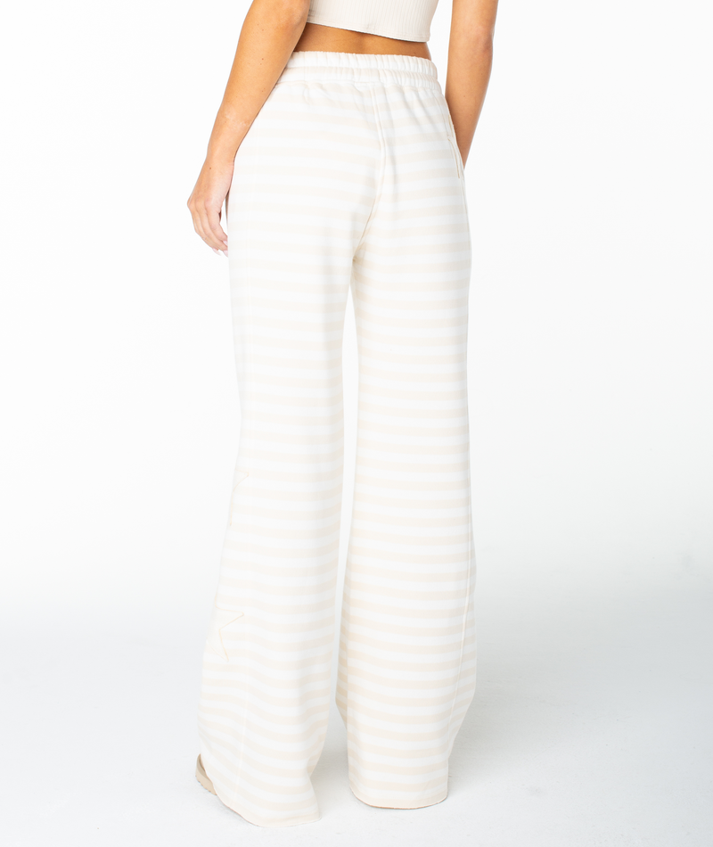 A person stands facing away with their arms relaxed by their sides, wearing the Roxy Fringing Reefs Pant—high-waisted, wide-leg trousers with cream and white stripes—set against a plain white background.